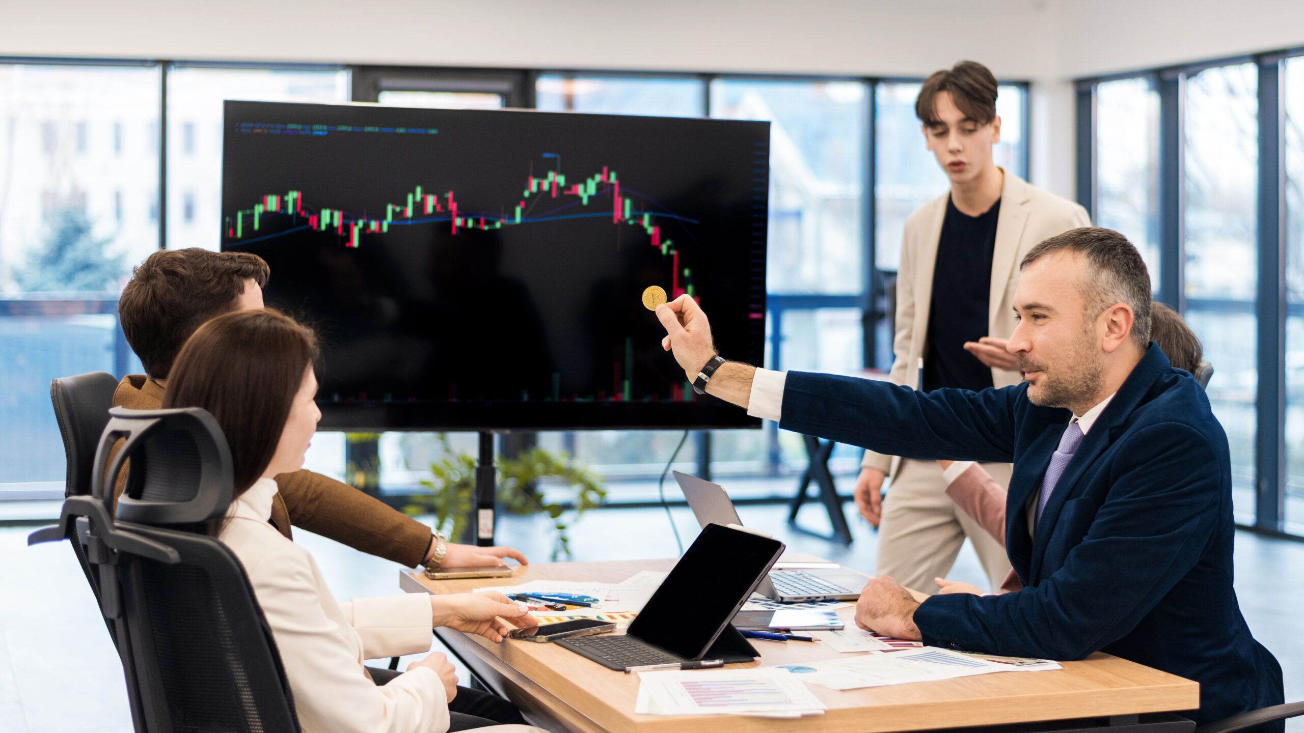 Young worker leading business meeting in an office, discussing the topic of cryptocurrencies with other workers using a big display with currency rate and physical coin